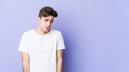 Young caucasian man isolated on purple background shrugs shoulders and open eyes confused.