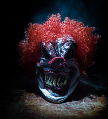 Spooky Clown head on wooden table. Evil clown head smiling on dark foggy background. Halloween...
