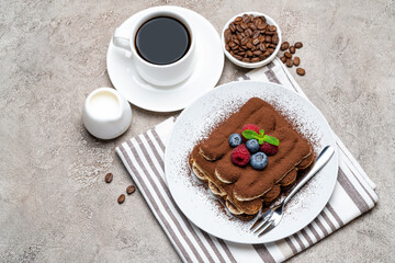 portion of Classic tiramisu dessert with raspberries and blueberries, cup of coffee and cream or milk on grey concrete background