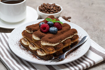 portion of Classic tiramisu dessert with raspberries and blueberries, cup of coffee and cream or milk on grey concrete background