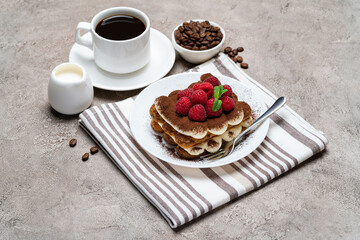 portion of Classic tiramisu dessert with raspberries and cup of coffee and cream or milk on grey concrete background