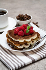 portion of Classic tiramisu dessert with raspberries and cup of coffee and cream or milk on grey concrete background