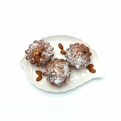 Three raisin muffins lie on a white leaf-shaped dish. Dusted with powdered sugar. Nearby are several almond kernels. Isolated over white background.