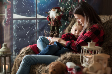 Fototapeta na wymiar Beautiful young mother, hugging her toddler boy, sitting in cozy chair on Christmas, decoration around