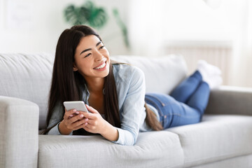 Carefree Asian Lady Enjoying Rest On Comfortable Sofa With Smartphone, Sincerely Smiling