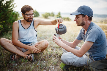 Outdoors image of young explorer man drinking hot beverage in mountains, sitting and relaxing after trekking.