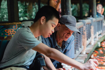 Fototapeta premium Asian disabled child on wheelchair spend holidays doing outdoor activities with fun, A boy feeding colorful japan kraft fish, Life in the education age of special Children, Happy disabled kid concept.
