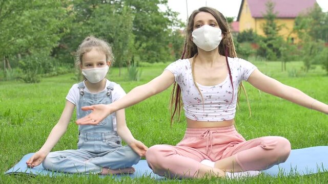 Young mother with dreadlocks and little daughter are doing yoga exercises on grass in the park wearing textile face mask due to quarantine. Summer vacation during lockdown concept
