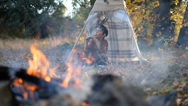 A little boy dressed in a Native American costume, sat down by the wigwam and played the flute. Playing with Indians near the fire with feathers on his head. Native American Heritage Day