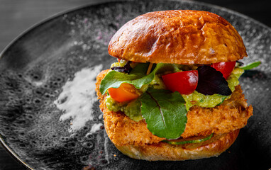 fish burger with cheese, guacamole sauce and vegetables in plate on black wooden table background