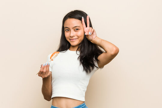 Young asian woman holding a sushi piece showing victory sign and smiling broadly.
