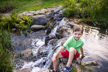 A young boy sits on the river bank, view from top. Warm summer or spring day. Cute Caucasian kid play with water