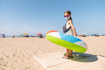 girl with a mask on her face and glasses walking down a path to the beach with a colorful inflatable mattress wheel on her arms to enjoy a vacation in the midst of the covid 19 coronavirus pandemic