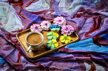 A Cup of black coffee with cookies decorated with sugar flowers of different colors on a wooden rectangular plate. The plate on a decorative fabric. The only light source.
