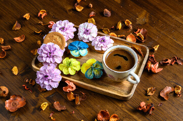 A Cup of coffee and cookies decorated with sugar flowers in different colors on a wooden rectangular plate. Dried fruits are scattered around on the wooden tabletop. A single light source.