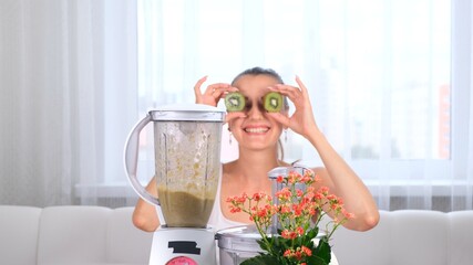 happy young woman preparing delicious nutritious smoothie in blender. funny female making eyes from kiwi.