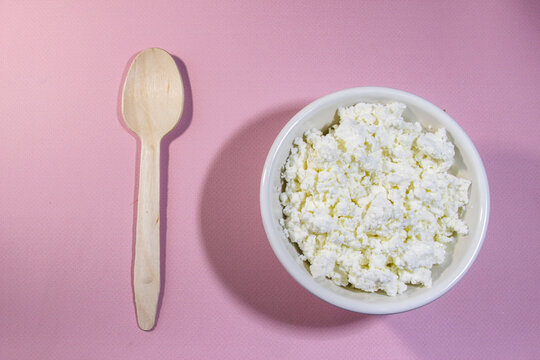 Cottage cheese in white bowl on pink table with spoon. Top view.