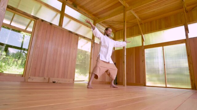 A Caucasian bearded man with long blond hair practices qigong and taichi in a wooden practice room in the summer forest. The practice of martial arts in European territories