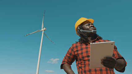 Adult or mature professional african man in hard hat standing with modern digital tablet against windmill power station making inspections of objects. High quality photo