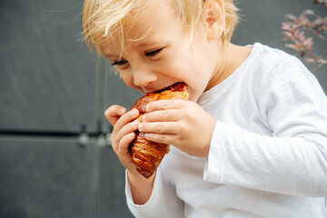 gluttonous little boy with blond hair eating crispy croissant outdoors on street