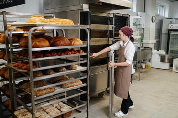 Working female baker standing next to a iron tray shelf with fresh bread