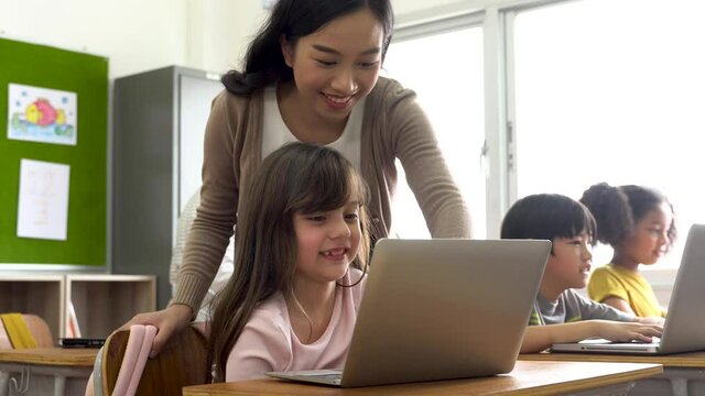 Young Asian teacher watching Caucasian school girl using computer in school classroom, digital native, technology, learning, guidance, education. Female instructor and student smiling.
