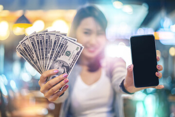 Young woman showing her mobile phone with a bunch of dollar banknotes. Make money online with smartphone concept.