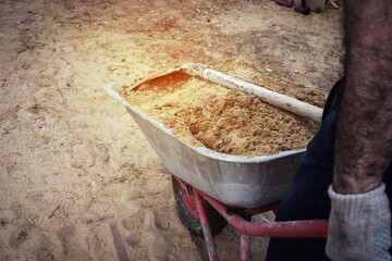 The gardener is driving a wheelbarrow with a shovel and filled with sand. Rear view