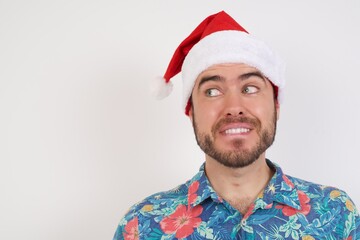 Photo of amazed Young caucasian man wearing hawaiian shirt and Santa hat over isolated white background bitting lip and looking up to empty space,