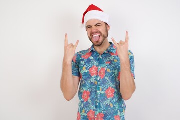 Young caucasian man wearing hawaiian shirt and Santa hat over isolated white background showing rock and roll hand gesture