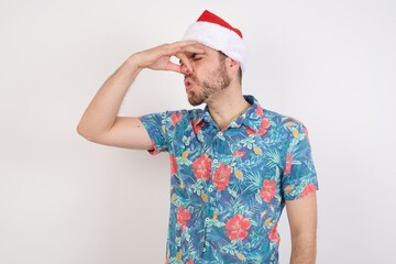 Young caucasian man wearing hawaiian shirt and Santa hat over isolated white background smelling something stinky and disgusting, intolerable smell, holding breath with fingers on nose. Bad smell
