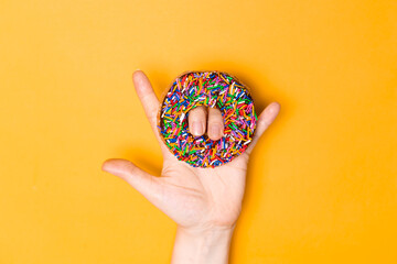 I love you hand sign with chocolate frosted donut with sprinkles, sugar-glazed frosted on yellow background. Playful and joyful tasty sugary comfort food for customer with copy space. Donut lovers.