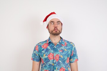 Young caucasian man wearing hawaiian shirt and Santa hat over isolated white background making fish face with lips, crazy and comical gesture. Funny expression.