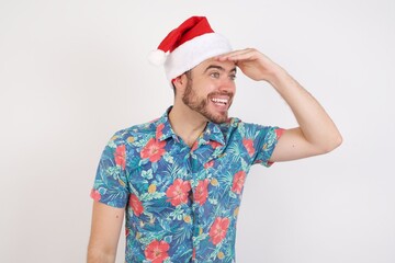 Young caucasian man wearing hawaiian shirt and Santa hat over isolated white background very happy and smiling looking far away with hand over head. Searching concept.
