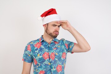 Young caucasian man wearing hawaiian shirt and Santa hat over isolated white background having problems, worried and stressed holds hand on forehead.