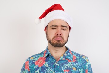 Dismal gloomy rejected Young caucasian man wearing hawaiian shirt and Santa hat over isolated white wall has problems and difficulties, curves lower lip and closes eyes in despair,