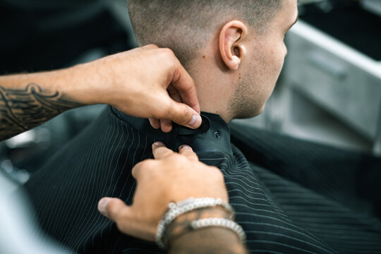 Cropped view of tattooed hairstylist wearing cape on man in barbershop 