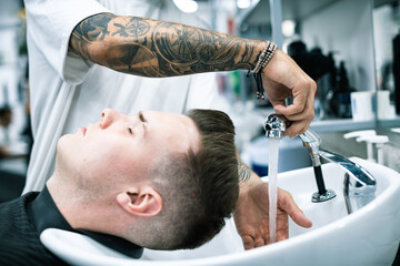 Selective focus of tattooed barber washing client hair before haircut in barbershop 