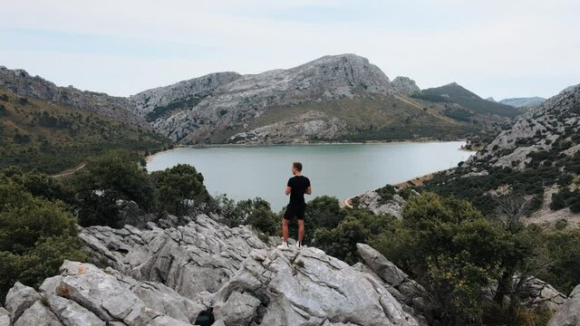 Aerial shot flying over a young man standing on a hill and revealing a mountain lake on background