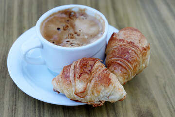 Cup of coffee and two croissants in a white saucer on wooden table. Tasty breakfast, cappuccino and fresh pastry close up