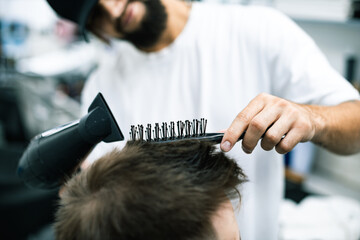 Cropped view of barber styling hair of young man with comb and dryer in barbershop 