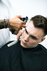 Tattooed barber holding hair clipper and comb near young man in cape 