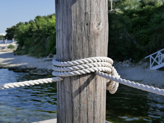 A thick white rope is attached to a massive wooden pole against the sea. Makeshift platform fence for sunbathing on the beach on a Sunny summer day.