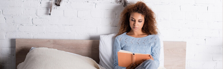 Panoramic shot of young woman in sweater reading book on bed