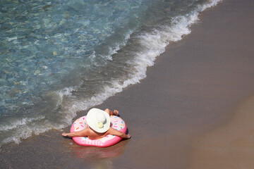 Woman in hat laying on inflatable donut ring on a sand in sea waves, aerial view. Beach vacation, relax and leisure in clean water