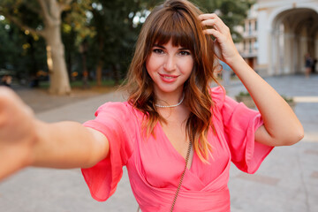 Elegant woman with perfect wavy hairs making self portrai by camera. Wearing pink dress.