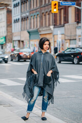Beautiful Latin young woman in grey stylish poncho and ripped torn jeans in urban street. Fashion and street style. Pretty hispanic brunette woman with short hair bob. Millennial in a city.
