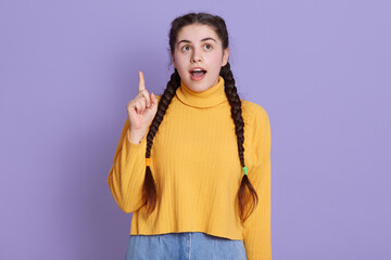 Pretty Caucasian woman with pensive and astonished expression, points with index finger up, dressed in yellow sweater, posing isolated over lilac background.