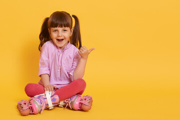 Happy girl in roller skating sitting on floor against yellow background showing thumbs aside with excited facial expression, copy space for advertisement or promotion.