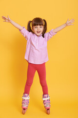 Happy little girl in shirt and leggins with roller skates posing indoors with hands up, standing against yellow background, cute kid looks excited and gladness.